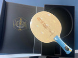 A table tennis paddle with a light wood blade and blue handle, displayed inside an open black presentation box next to the inner lid featuring gold branding.