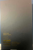 A black rectangular product box with gold text and barcodes on one side, photographed on a blue surface with part of a person's leg visible at the left edge.