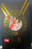 A black rectangular product box with gold and red printed graphics, photographed on a blue surface. The box features large gold text reading 'RIO TOKYO PARIS LONDON' around a circular team photo, a red square logo near the top left, and gold script text on the left side.